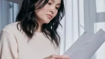 窓辺で書類を読む人、薄いセーターを着ている。
