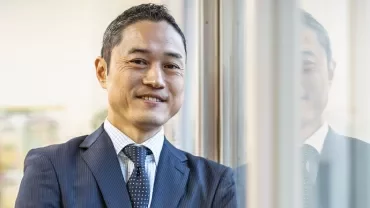 窓際に立つスーツ姿の笑顔の男性。