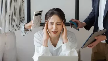 机の上でストレスを感じている女性と、書類を持つ二人。