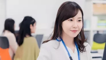 オフィスでコンピュータを使っている女性、白いジャケットと青いストラップを着用。