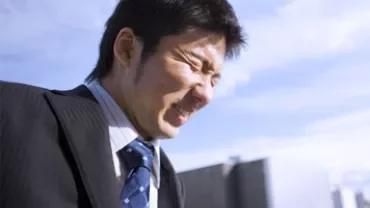 Man squinting in sunlight in front of buildings.
