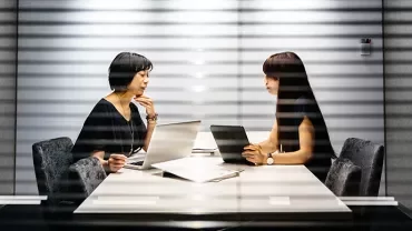 Two women discussing work at a conference table with laptops.
