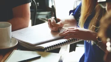 People writing in notebooks and drinking coffee at a table.