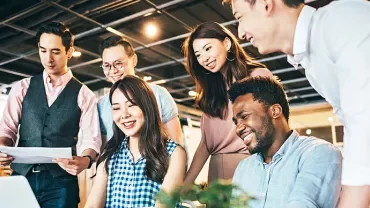 Group of coworkers gathered around a laptop, smiling and collaborating in an office.