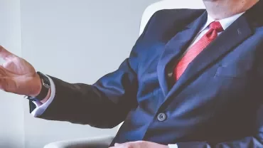Man in a suit with a red tie gesturing while seated.