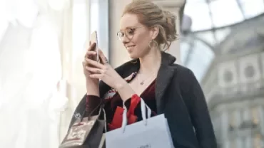 Woman smiling while looking at her phone and carrying shopping bags.