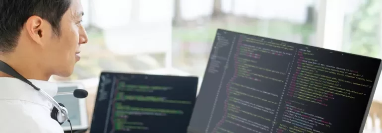 ヘッドセットを付けた人が、コードを表示したデュアルモニターで作業している。