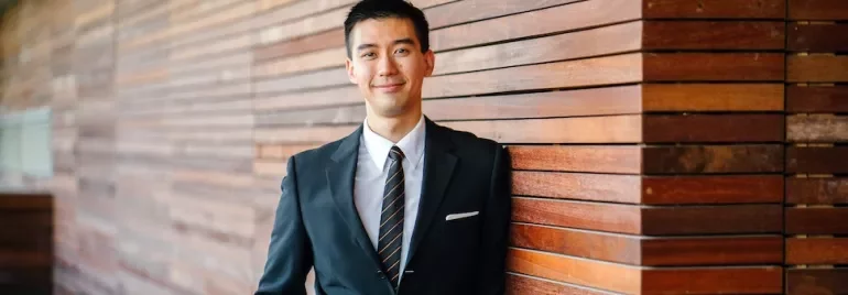 スーツ姿の男性が木の壁に寄りかかって微笑んでいる。