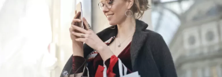 Woman smiling while looking at her phone and carrying shopping bags.
