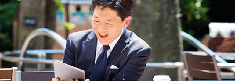 屋外でタブレットを見ながら笑っているスーツ姿の男性、テーブルにコーヒーカップ。