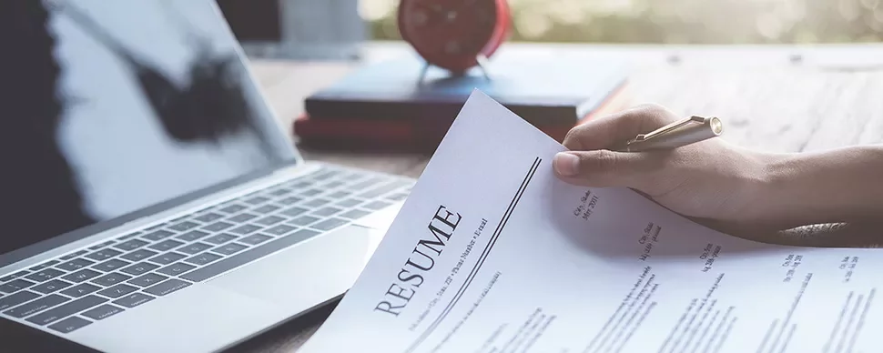 A person holding a resume in front of a laptop on a desk.