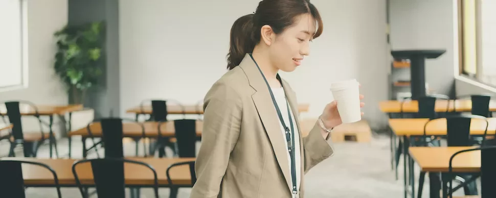 オフィスでコーヒーカップを持って歩く女性。背後にはテーブルと椅子が並んでいる。