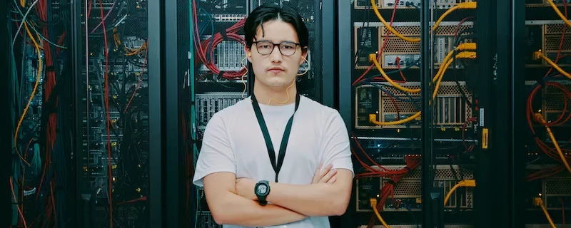 メガネをかけた白いシャツの人物がサーバールームに立っている。