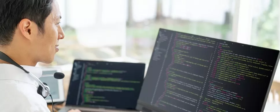 ヘッドセットを付けた人が、コードを表示したデュアルモニターで作業している。