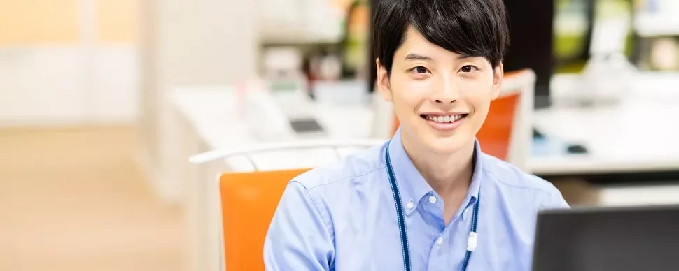 オフィスでノートパソコンを使って作業する青いシャツの笑顔の人物。