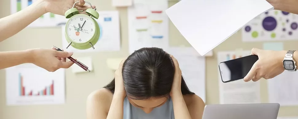 机に座って頭を抱える人物と周りの手が時計やペン、電話、紙を持っている。