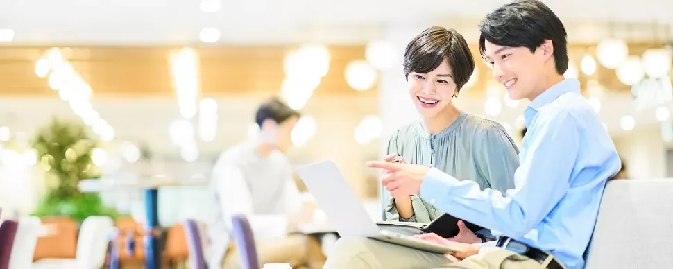 明るいオフィスで、ノートパソコンを見ながら笑顔の二人。