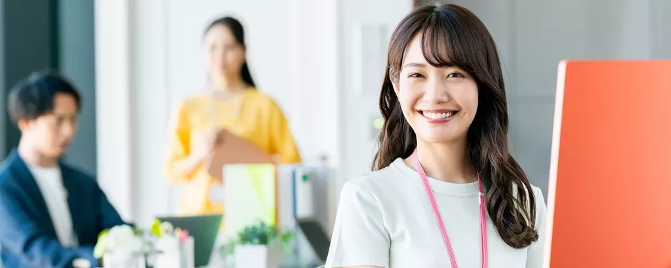 笑顔の女性がデスクで働いており、背景に同僚がいます。