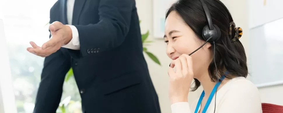 ヘッドセットを着けた女性が微笑んでおり、近くで人が指示を出しています。
