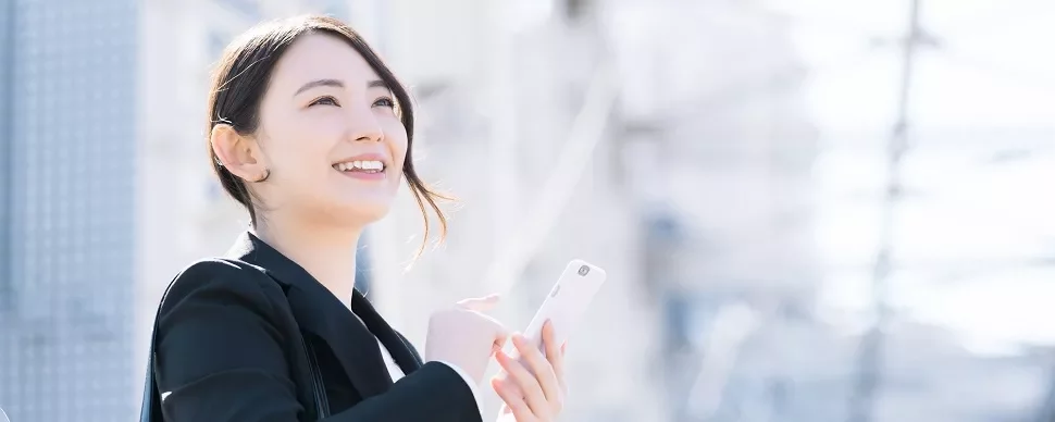 屋外で微笑んでスマートフォンを持つスーツ姿の人物。