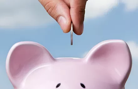 A hand inserting a coin into a pink piggy bank against a blue sky.