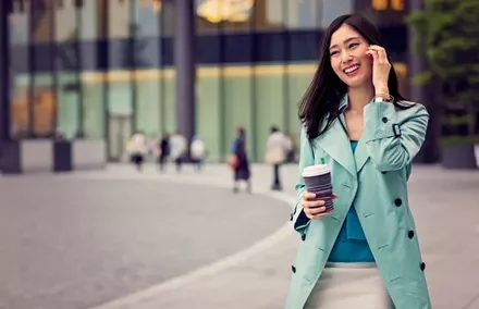 Woman in blue coat smiles while holding a coffee cup, walking outdoors.