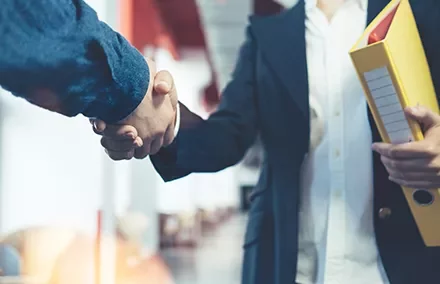 Two people shaking hands, one holding a yellow folder, in an office setting.
