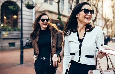 Two women walking in a city, smiling and wearing sunglasses.