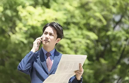 Man in a suit talks on phone while reading a newspaper outdoors.