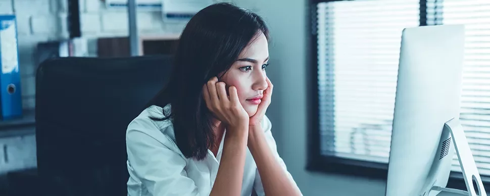 A woman in a white shirt is sitting at a desk, looking intently at a computer screen.