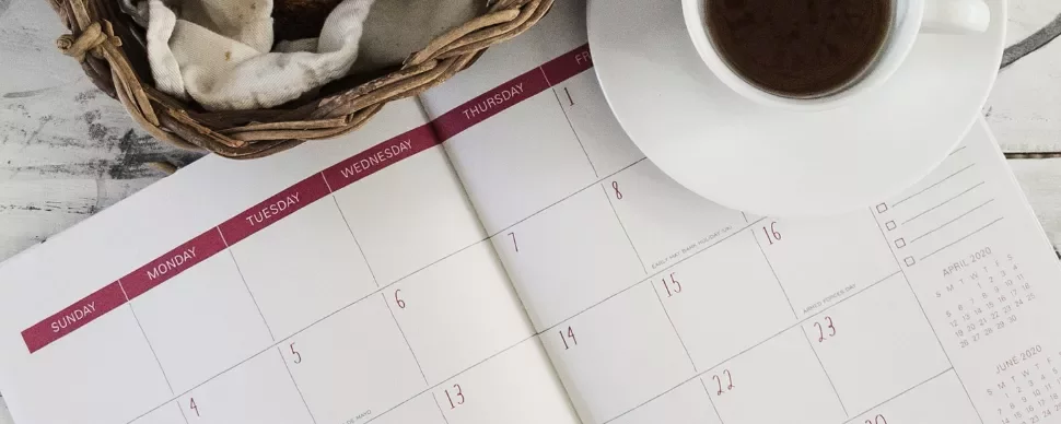 Open calendar with a cup of coffee and a bread basket on a wooden table.