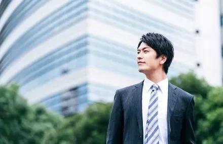 A man in a suit stands in front of a modern building, looking into the distance.