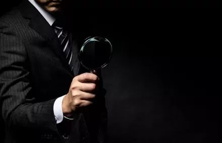 Person in a suit holding a magnifying glass against a dark background.