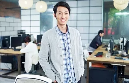 Man smiling in a modern office setting with colleagues working in the background.
