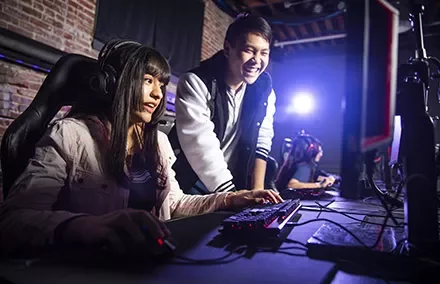 Two people gaming on computers in a dark room, one smiling and standing, the other seated and focused.