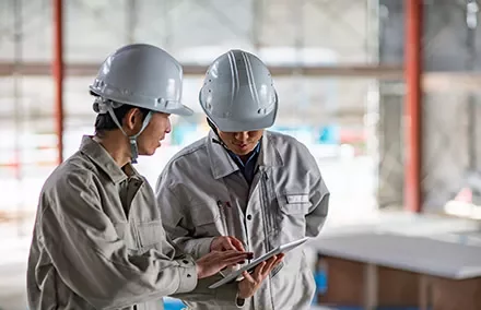 Two workers in helmets reviewing plans on a tablet at a construction site.