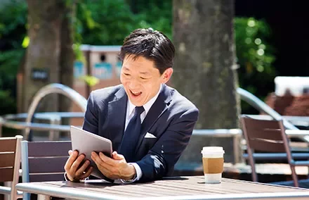 Smiling man in a suit looking at a tablet outdoors, with a coffee cup on the table.