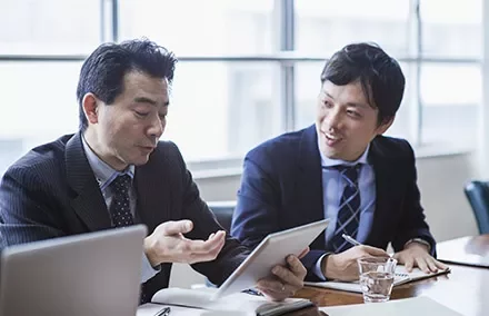 Two men in suits discussing with a tablet and notebook.