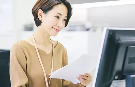 Woman at desk holding papers and smiling at computer.