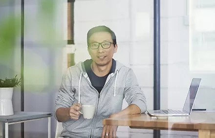フーディーを着た男性がコーヒーカップを持ち、ノートパソコンの横に座っている。